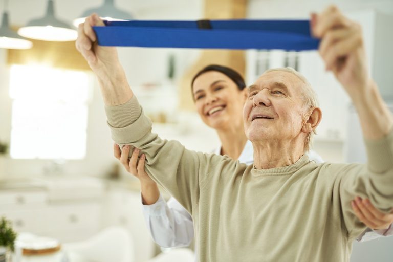 Happy young nurse helping an elderly man to do exercises in room